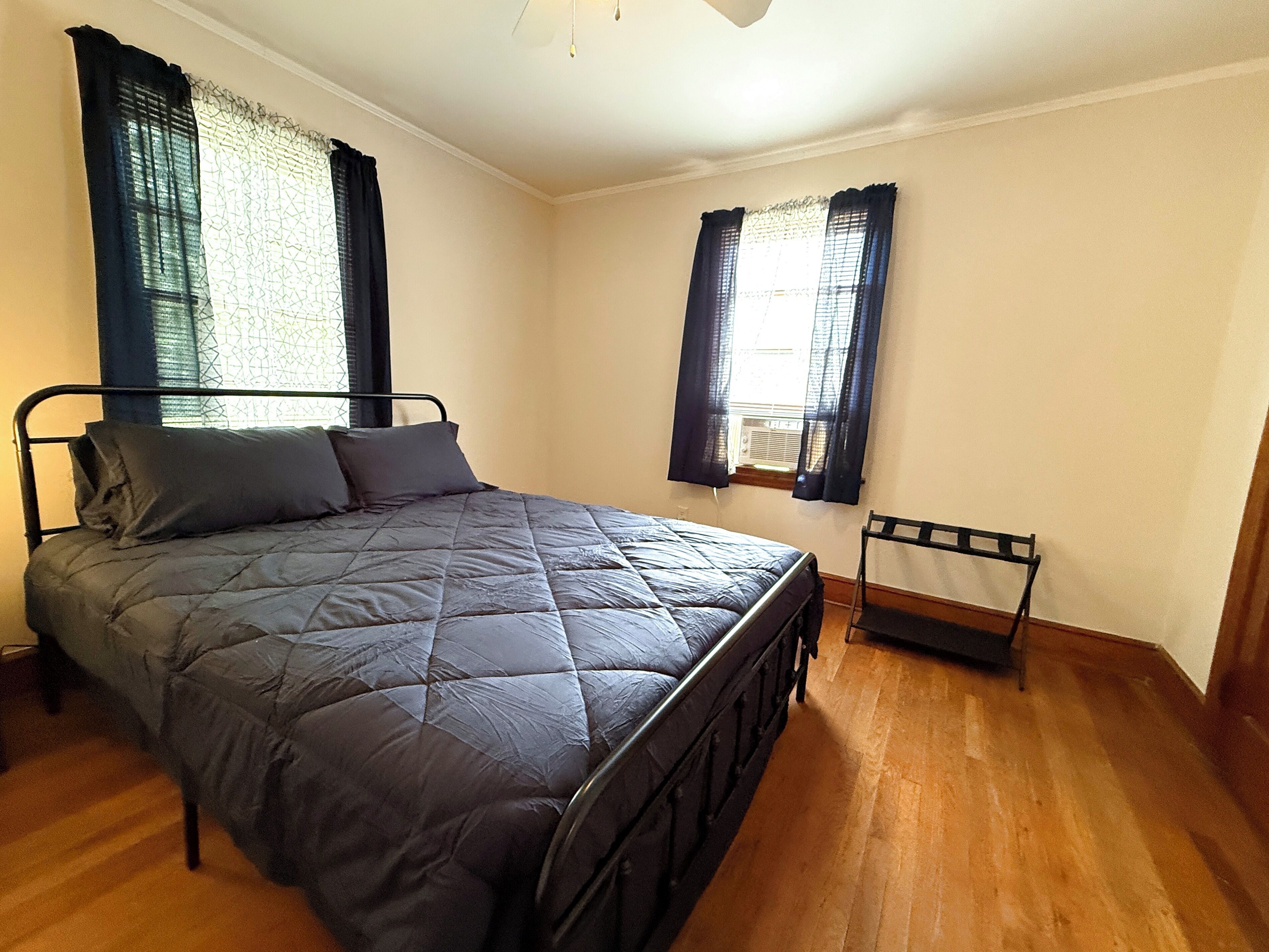 A bedroom with a bed, two windows with curtains, a radiator, and a wooden floor.