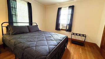 A bedroom with a bed, two windows with curtains, a radiator, and a wooden floor.