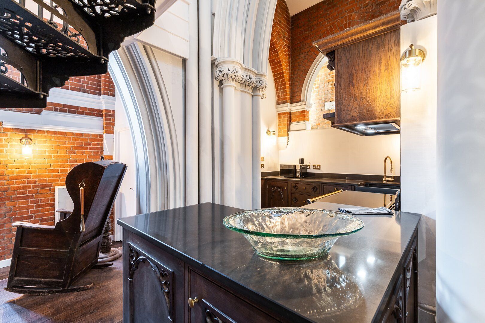 A kitchen with a glass bowl on the counter, a brick wall, and a wooden chair.