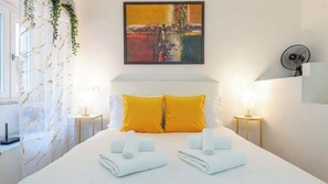 Ein Hotelzimmer mit einem Bett, zwei Kissen, einem Nachttisch, einem Wandbild, einem Standventilator und einem Fenster mit Vorhängen.