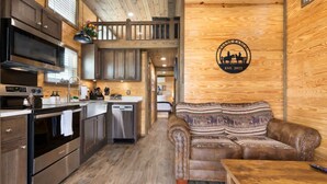 A modern kitchen with stainless steel appliances, wooden cabinets, and a dining area with a brown leather couch. - Tiny Home 140 - Evans Ranch (Fredericksburg)