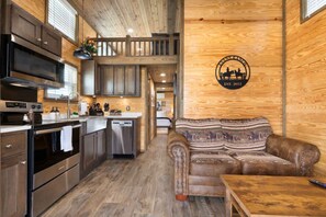 A modern kitchen with stainless steel appliances, wooden cabinets, and a dining area with a brown leather couch. - Tiny Home 140 - Evans Ranch (Fredericksburg)