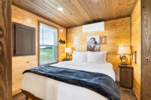 A wooden-paneled bedroom with a bed, two bedside lamps, a window with blinds, and a wall-mounted air conditioning unit. - Evans Ranch | Rooftop Deck Views (Fredericksburg)