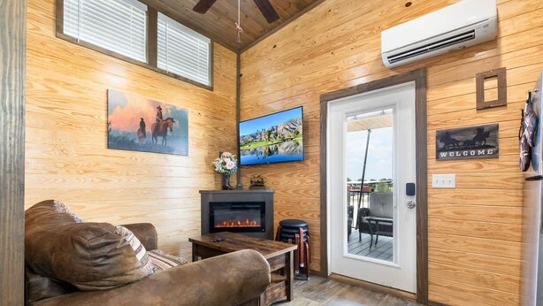 A cozy living room with a fireplace, wooden walls, a television, and a door leading outside. - Tiny Home 140 - Evans Ranch (Fredericksburg)