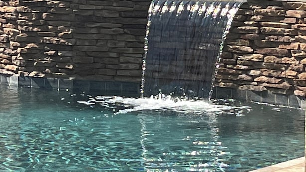 Een zwembad met waterval, omringd door een stenen muur en een metalen hek.