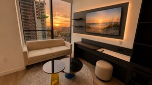 Ein modernes Wohnzimmer mit einer Couch, einem an der Wand montierten Fernseher und Blick auf einen Sonnenuntergang durch ein großes Fenster.