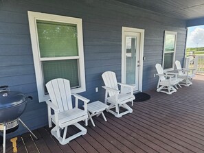 Eine überdachte Veranda mit weißen Adirondack-Stühlen, einem Grill und einer Tür mit Fenster.