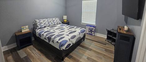 Quarto com cama, ventilador de teto, mesa de cabeceira, luminária, janela com persianas e televisão fixada na parede.