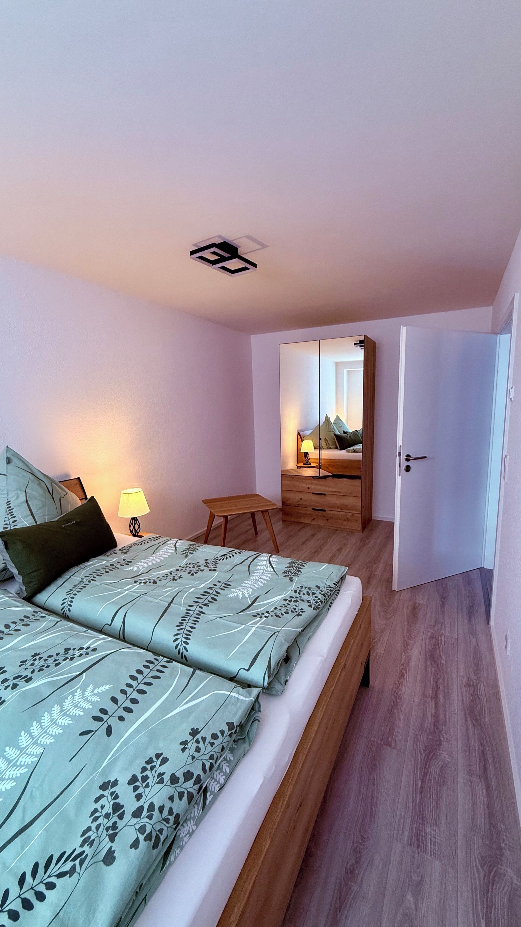Una camera da letto con un letto, una lampada da comodino, un comodino in legno e vista su un'altra stanza attraverso una porta aperta.