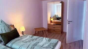 Una camera da letto con un letto, una lampada da comodino, un comodino in legno e vista su un'altra stanza attraverso una porta aperta.