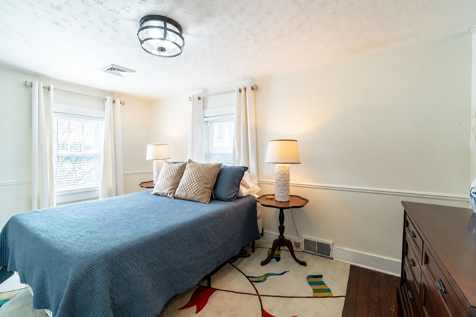 Une chambre à coucher avec un lit, deux tables de chevet avec des lampes, une commode et une fenêtre avec des rideaux.