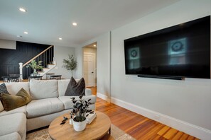 A modern living room with a large flat-screen TV, a sofa, a coffee table with decorative items, and a staircase leading to another level. - 5 Mi to Boston & Airport! Townhome w/ Skyline View (Everett)