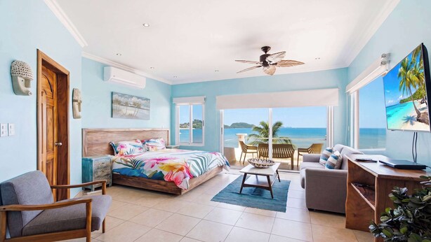 A bedroom with a bed, a desk, a chair, a TV, and a balcony with a view of the sea. - Beachfront Penthouse with Panoramic Ocean Views (Playa Potrero)