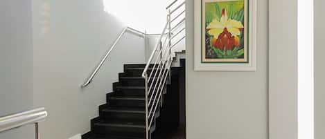 Un escalier avec une rampe métallique, un palier recouvert d’une moquette noire et un tableau encadré représentant des fleurs, accroché au mur.
