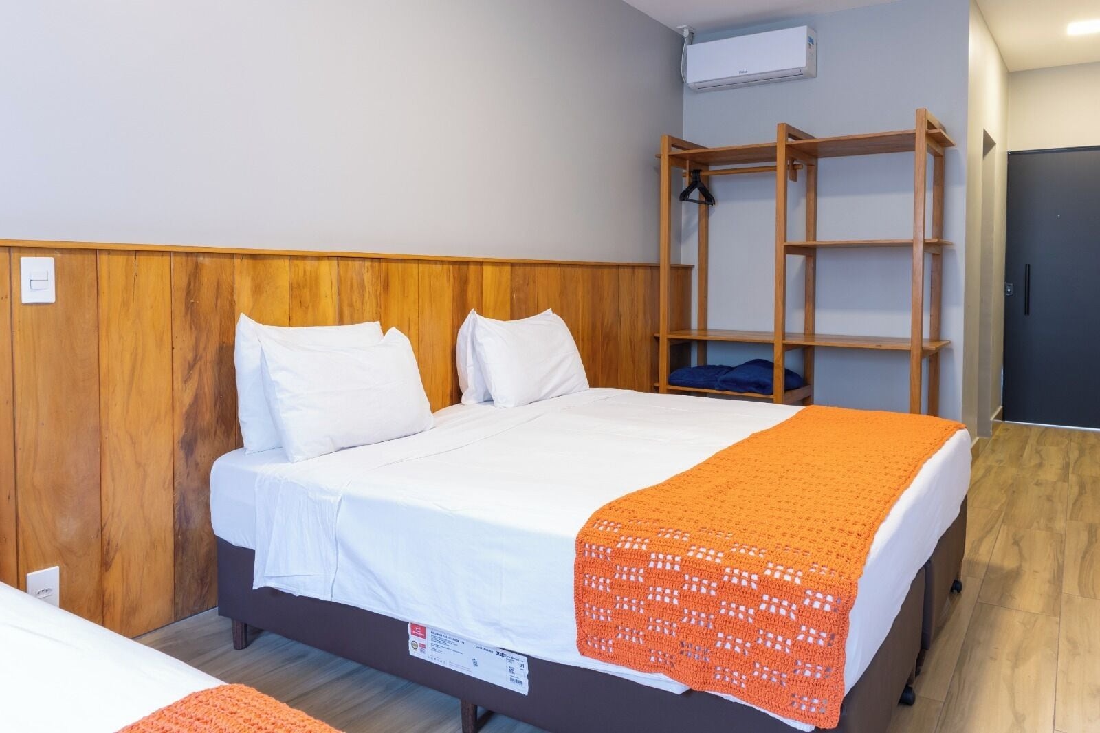 A single bed with a white mattress and an orange blanket, wooden headboard, and a small shelf unit with a hanging item.