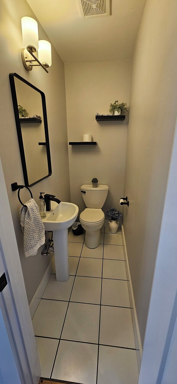 Une petite salle de bain avec un lavabo, des toilettes et un miroir, le tout de couleur blanche. On y trouve deux appliques murales et une petite étagère sur laquelle est posée une plante.