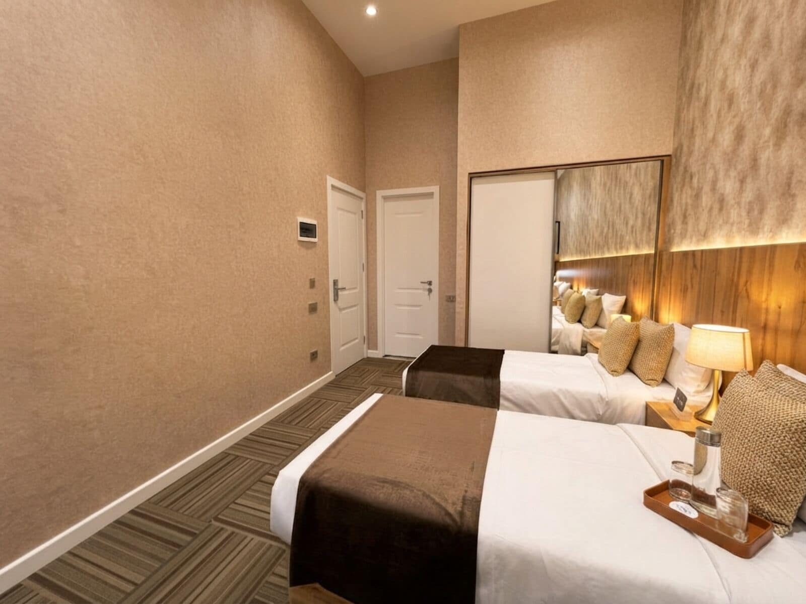 A hotel room with two beds, a large mirror, a nightstand with a lamp, and a tray with glasses.
