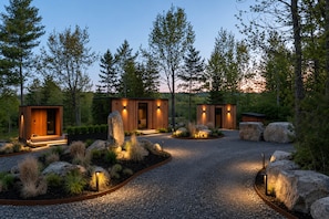 A modern outdoor cabin with a gravel driveway, surrounded by large rocks and a variety of plants.