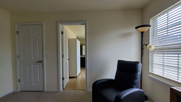 A room with a black chair, a door, a hallway, and a window with blinds.