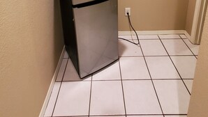 A small, single-door refrigerator with a power cord plugged in, located in a corner of a room with beige walls and white tiled flooring.