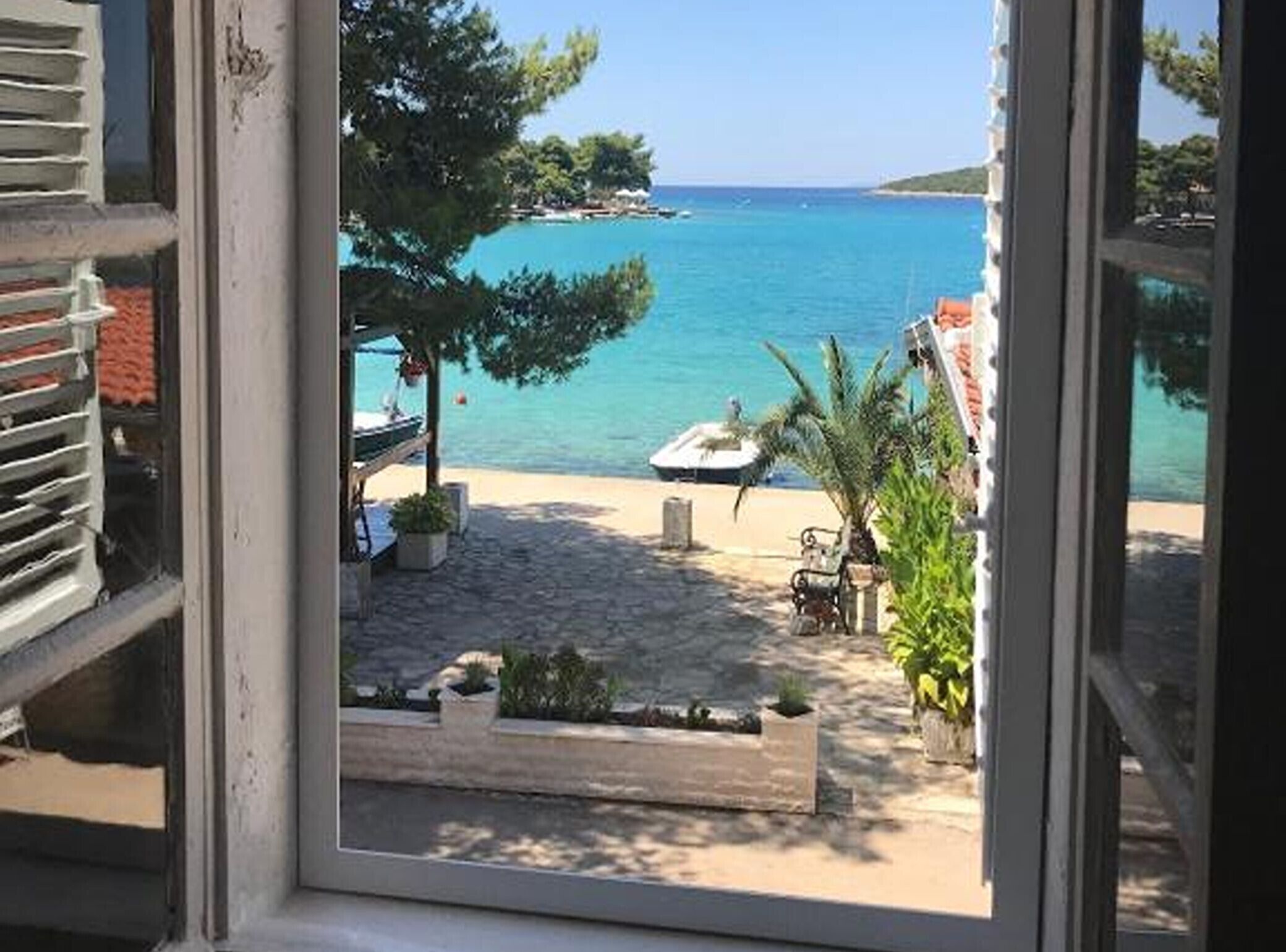 A view through a window of a beachside area with a paved walkway, outdoor seating, and a boat in the water.