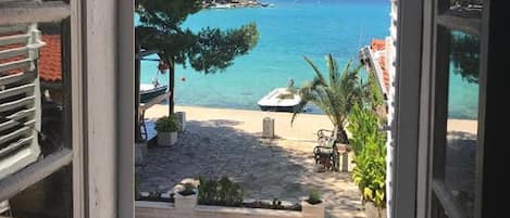 A view through a window of a beachside area with a paved walkway, outdoor seating, and a boat in the water.