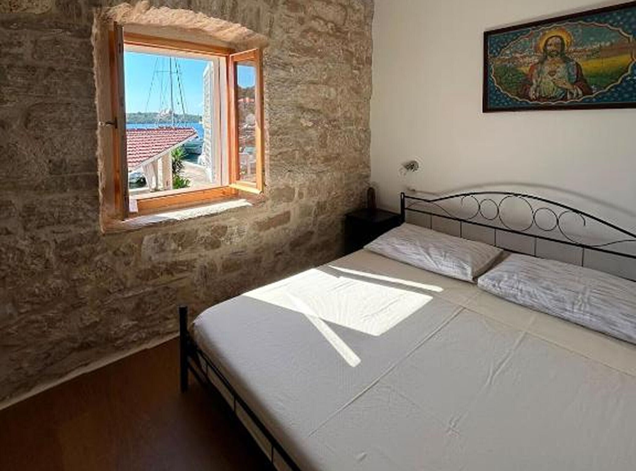 Een slaapkamer met een stenen muur, een raam met uitzicht op boten, een bed met witte lakens, en een schilderij aan de wand.