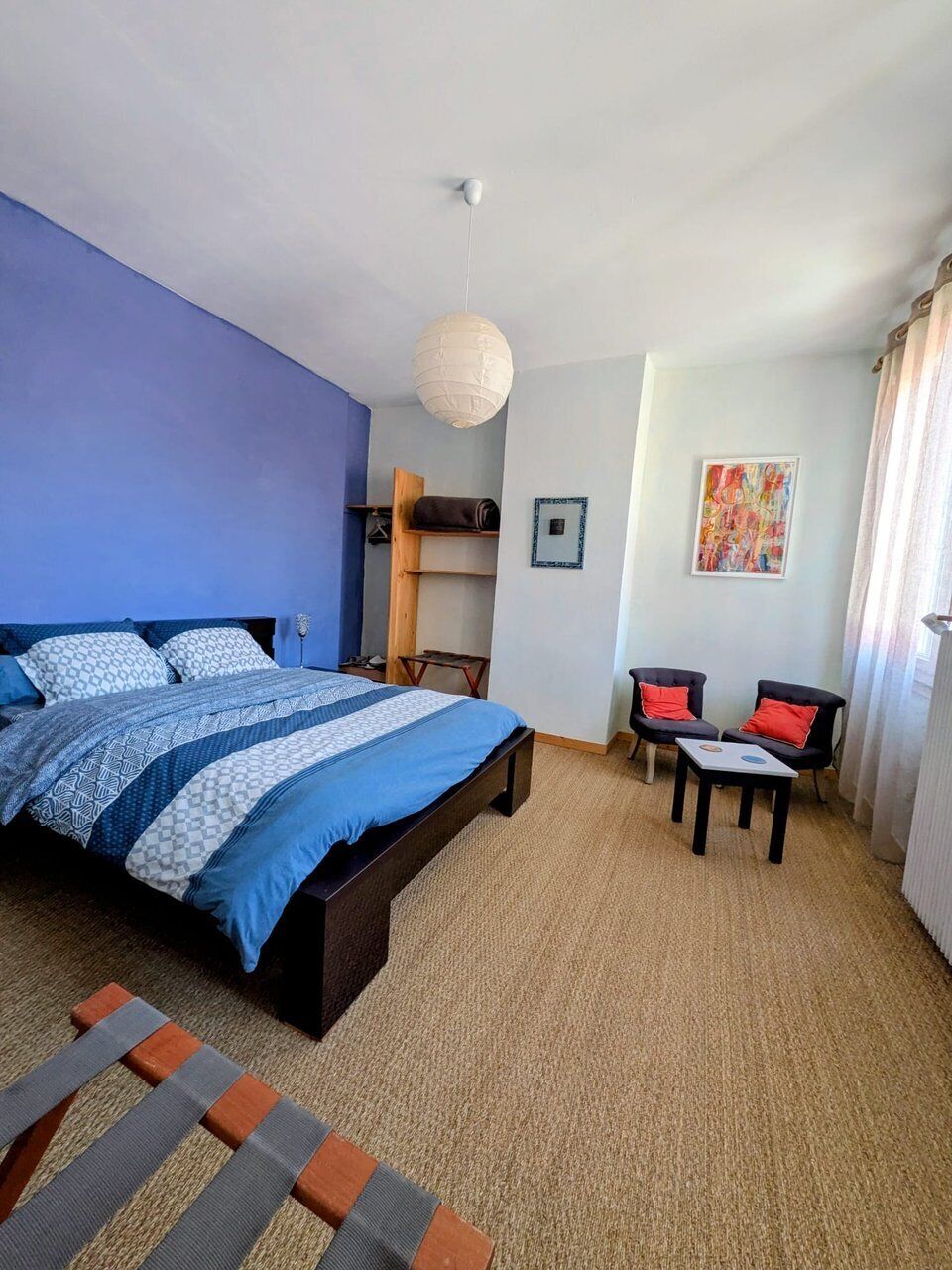 A bedroom with a bed, two chairs, a hanging light fixture, a framed picture, and a window with curtains.