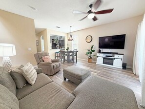 Ein Wohnzimmer mit Sofa, Hocker, Deckenventilator, Fernseher und einem Essbereich mit Stühlen und Tisch.