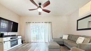 Un soggiorno con una TV a schermo piatto, un ventilatore a soffitto, un divano, un poggiapiedi e un angolo cottura con forno a microonde.