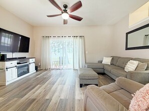 Ein Wohnzimmer mit Flachbildfernseher, Deckenventilator, einer Couch, einem Hocker und einer Küchenzeile inklusive Mikrowelle.