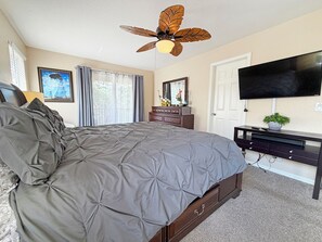 Ein Schlafzimmer mit Bett, Deckenventilator, Kommode, Fernseher und einem Fenster mit Vorhängen.