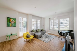 Una sala de estar moderna con un sofá seccional, un sillón amarillo, una mesa de comedor con sillas y ventanas grandes que ofrecen vistas de la ciudad.