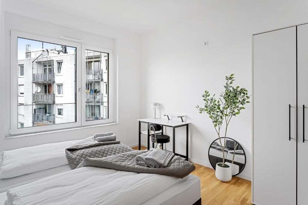 A modern bedroom with two beds, a desk, a chair, a potted plant, and a view of a building outside.