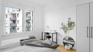A modern bedroom with two beds, a desk, a chair, a potted plant, and a view of a building outside.