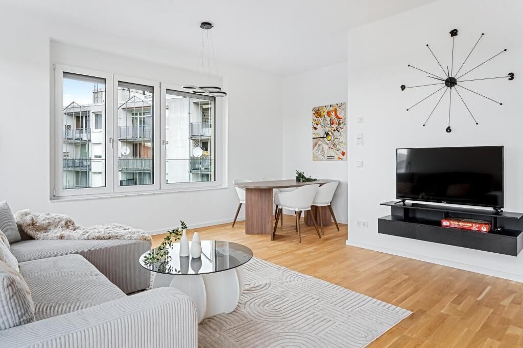 Un soggiorno moderno con un divano, un tavolino in vetro, un'area pranzo con tavolo e sedie in legno, una TV a parete e ampie finestre con vista sugli edifici.
