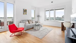 A modern living room with a sofa, a red chair, a round coffee table, and a view of the city through large windows. - EXQUIS 3 Room Serviced Apartment (Böblingen)