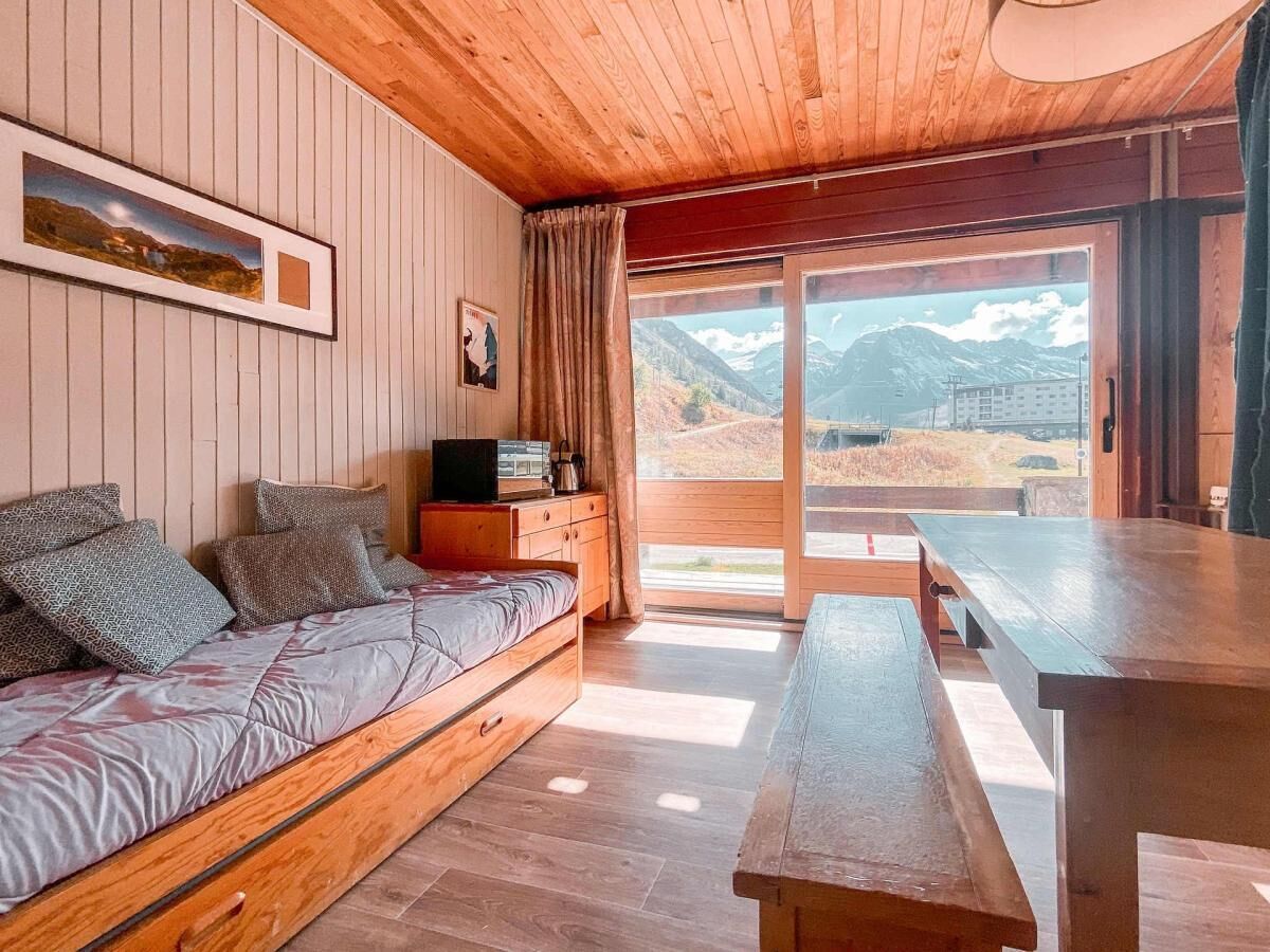 A cozy room with a wooden ceiling, a bed with patterned bedding, a small table, and a view of mountains through a large window.