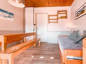 Una habitación acogedora con un banco de madera, una cama con almohadones estampados, un estante de madera con botellas, y una puerta.