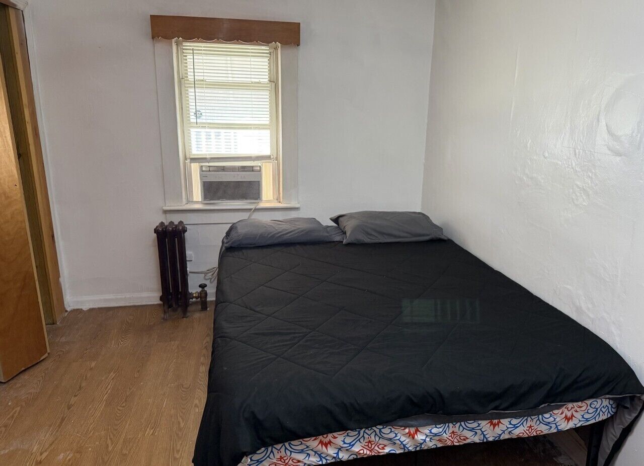 Ein kleines Zimmer mit einem Einzelbett, Holzboden, Heizkörper und einem Fenster mit Jalousien.