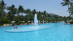 Una piscina grande con una fuente central, rodeada de palmeras y sillas de descanso.