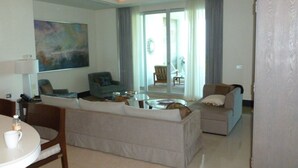 Una sala de estar con un sofá, un sillón y una mesa de comedor con sillas. Hay un ventanal con cortinas y vistas al exterior.