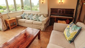A living room with a sofa, a coffee table, a piano, and a window with a view of greenery. - Claybottom Farm (Wigton)
