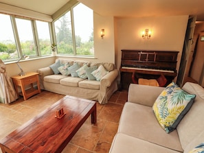 A living room with a sofa, a coffee table, a piano, and a window with a view of greenery. - Claybottom Farm (Wigton)