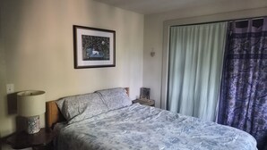 Quarto com cama, mesa de cabeceira, luminária, quadro na parede e janela com cortinas.