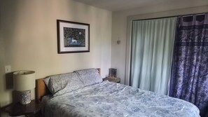 Ein Schlafzimmer mit einem Bett, einem Nachttisch, einer Lampe, einem gerahmten Bild an der Wand und einem Fenster mit Vorhängen.