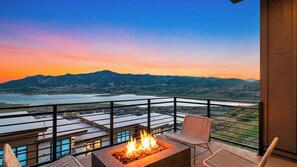 Un balcón con una chimenea, sillas de mimbre y vistas a montañas y un lago al atardecer.