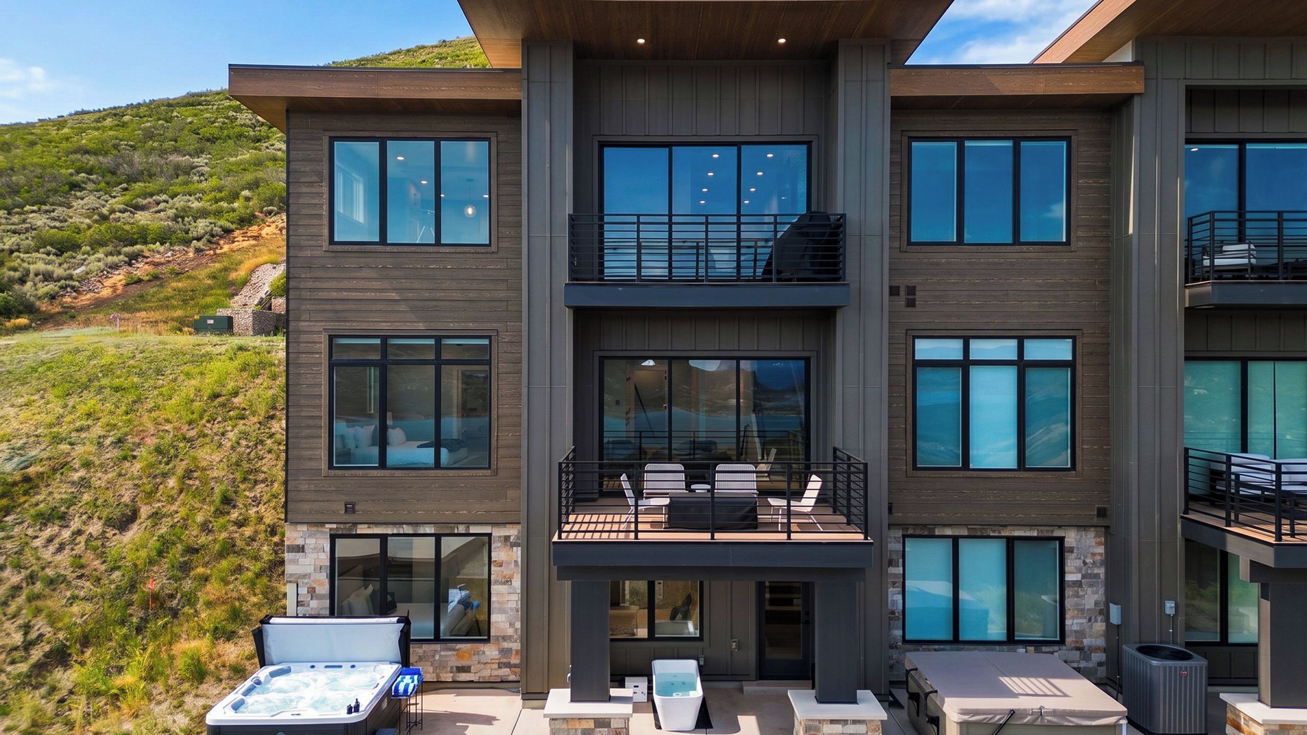 Un edificio moderno de varios pisos con grandes ventanales, un balcón con mobiliario exterior y un jacuzzi.