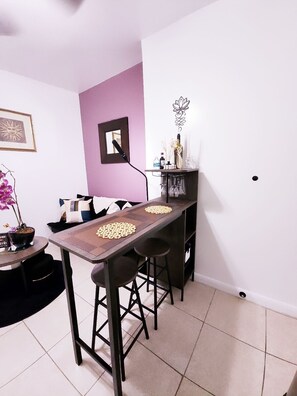 Un comedor con una mesa larga, dos taburetes de bar, una mesita auxiliar con una planta en maceta y un espejo montado en la pared.