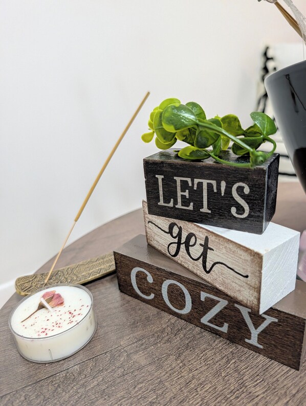 Ein Holzbruchstück mit dem Aufdruck 'LET'S GET COZY', eine kleine Kerze mit Docht und eine grüne Pflanze darauf.